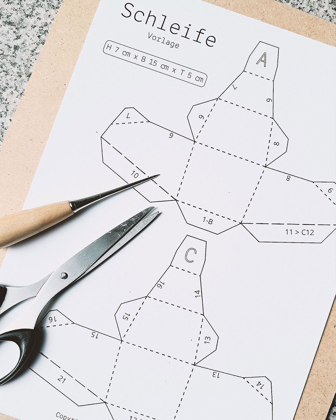 Ausgedruckte Bastelvorlage (PDF/SVG) für eine Low-Poly Papierschleife, mit Schere und Bleistift auf Unterlage