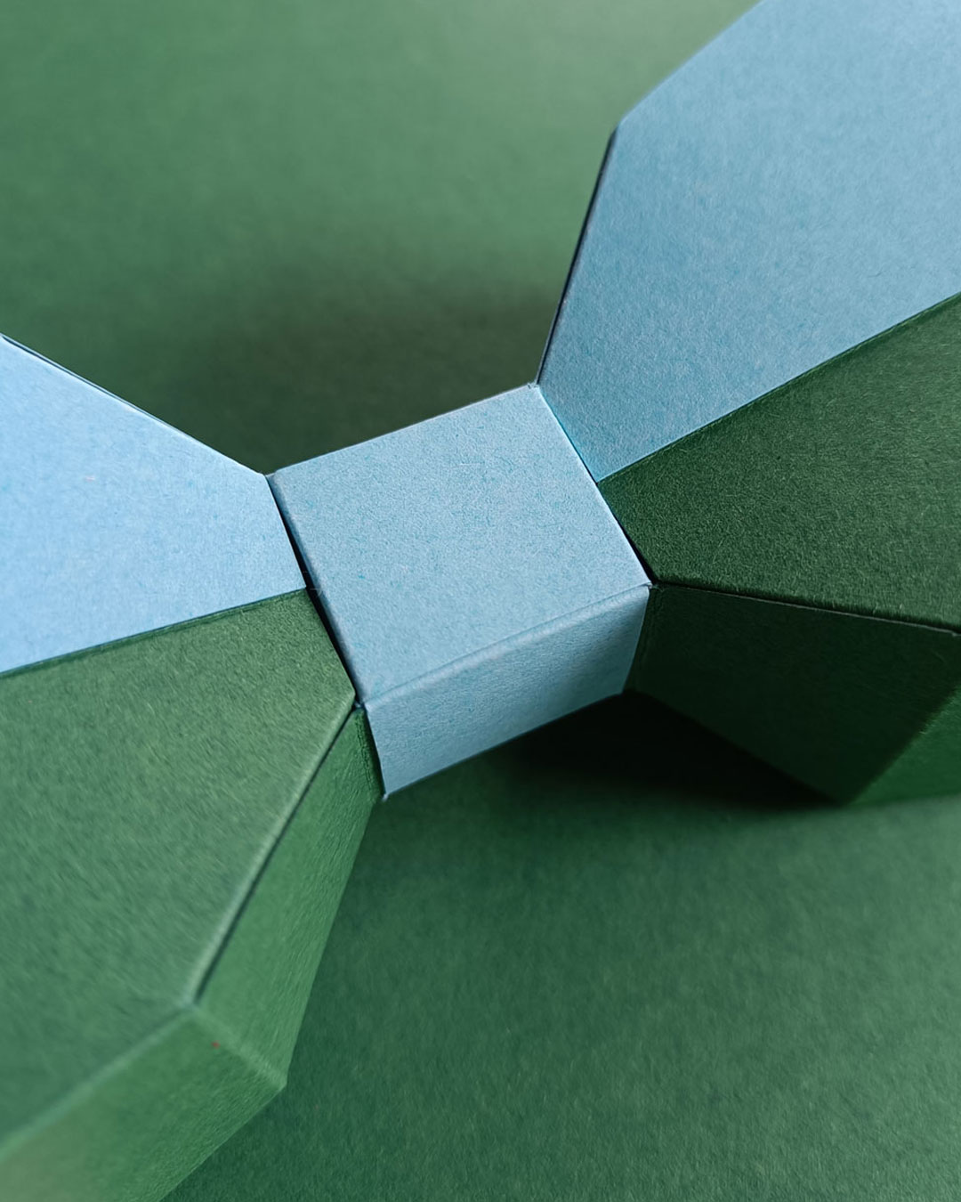 Makroaufnahme einer 3D Low-Poly Papierschleife, Fokus auf Papierfasern und scharfe Kanten in Grün- und Blautönen.