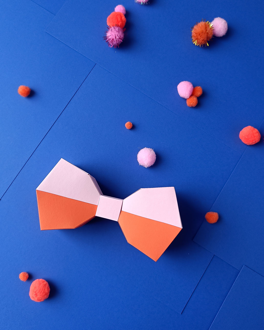3D Low-Poly Papierschleife in Orange und Puderrosa auf tiefblauem Hintergrund mit bunten Pompons - starker Kontrast