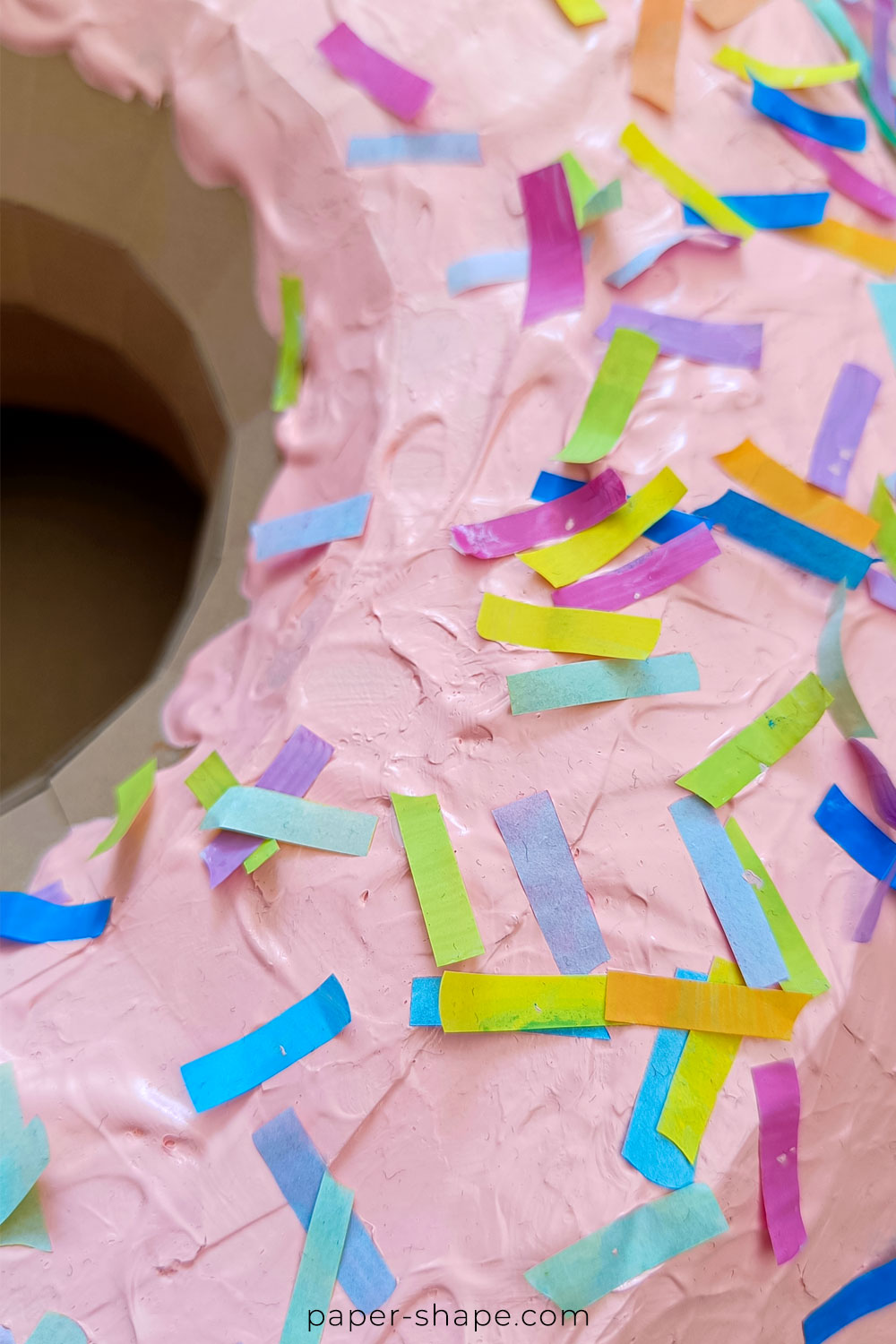 Detailaufnahme: Bunte Papierstreifen als Streusel auf pink bemalter Donutoberfläche aus recyceltem Material.